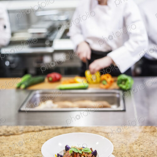 A closeup of a finished plate of food with chefs working in the background in a… A closeup of a finished plate of food with chefs working in the background in a…