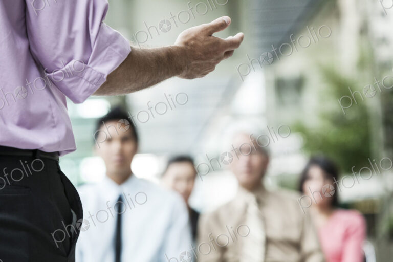 A closeup of a hand gesturing to a group of business people in a meeting.