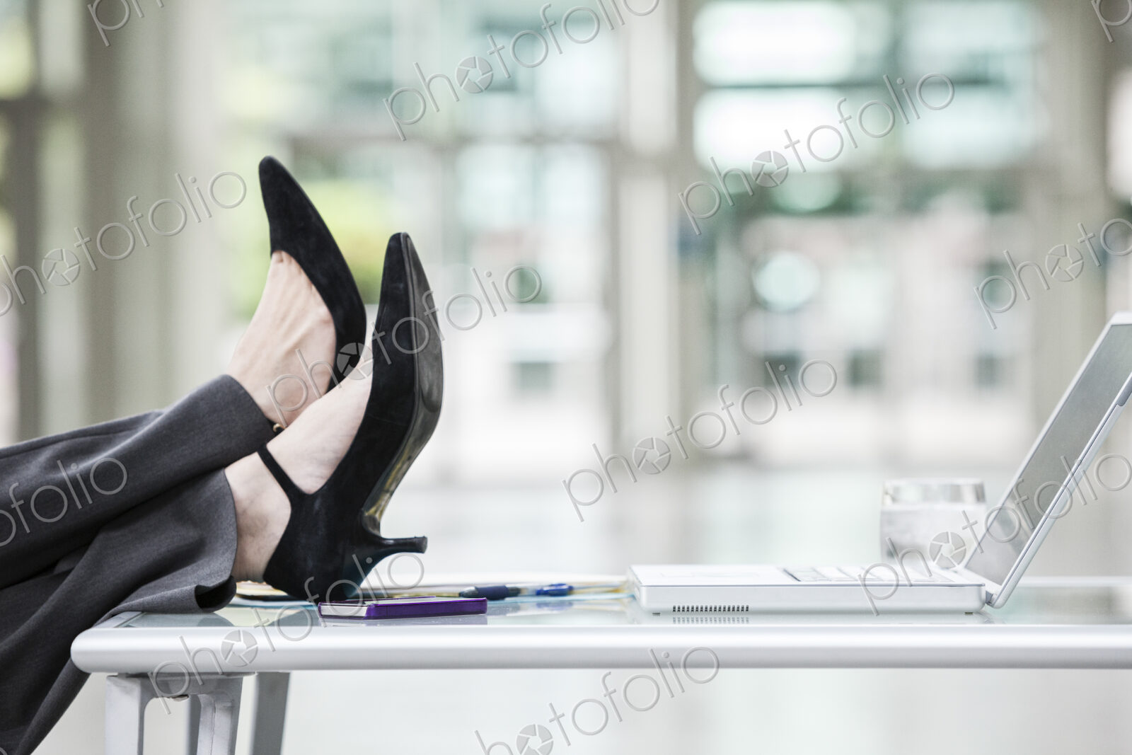 A closeup of feet and high heel shoes resting on a desk in front of a laptop computer.