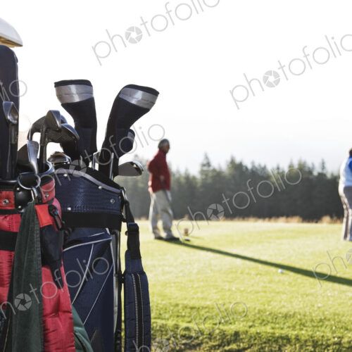 A closeup of golf clubs on a cart with golfers on the green in the background.
