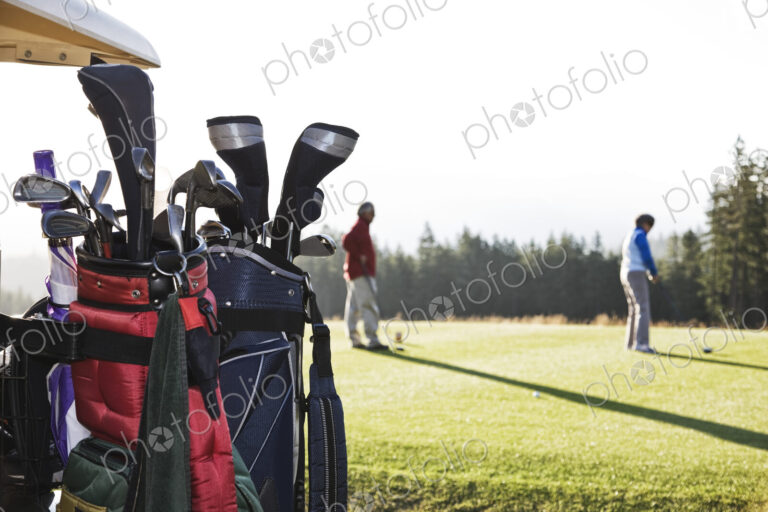 A closeup of golf clubs on a cart with golfers on the green in the background.