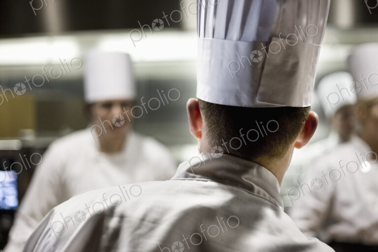 A closeup view from behind of a chef wearing a toque hat in a commercial kitchen.