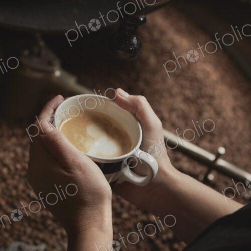 A coffee shop. A person holding a fresh brewed cup above a metal drum with roasting…
