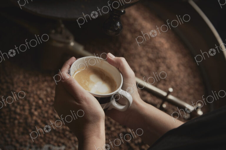 A coffee shop. A person holding a fresh brewed cup above a metal drum with roasting…