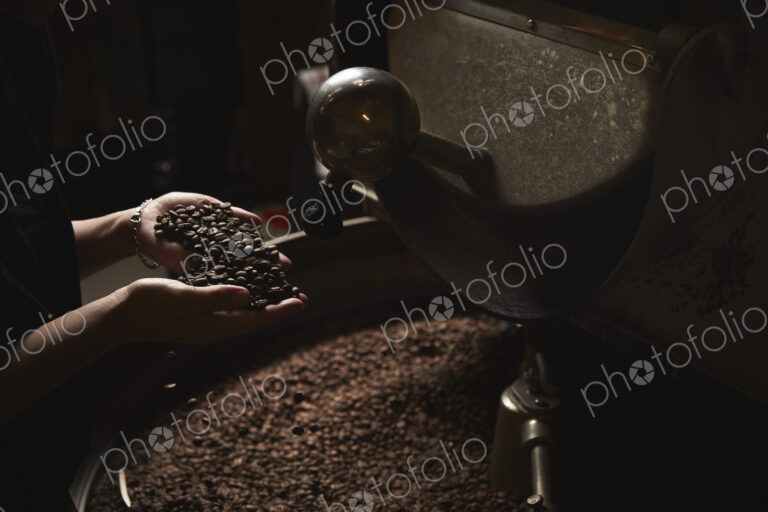 A coffee shop. A person holding a handful of fresh roasted beans above a metal drum…