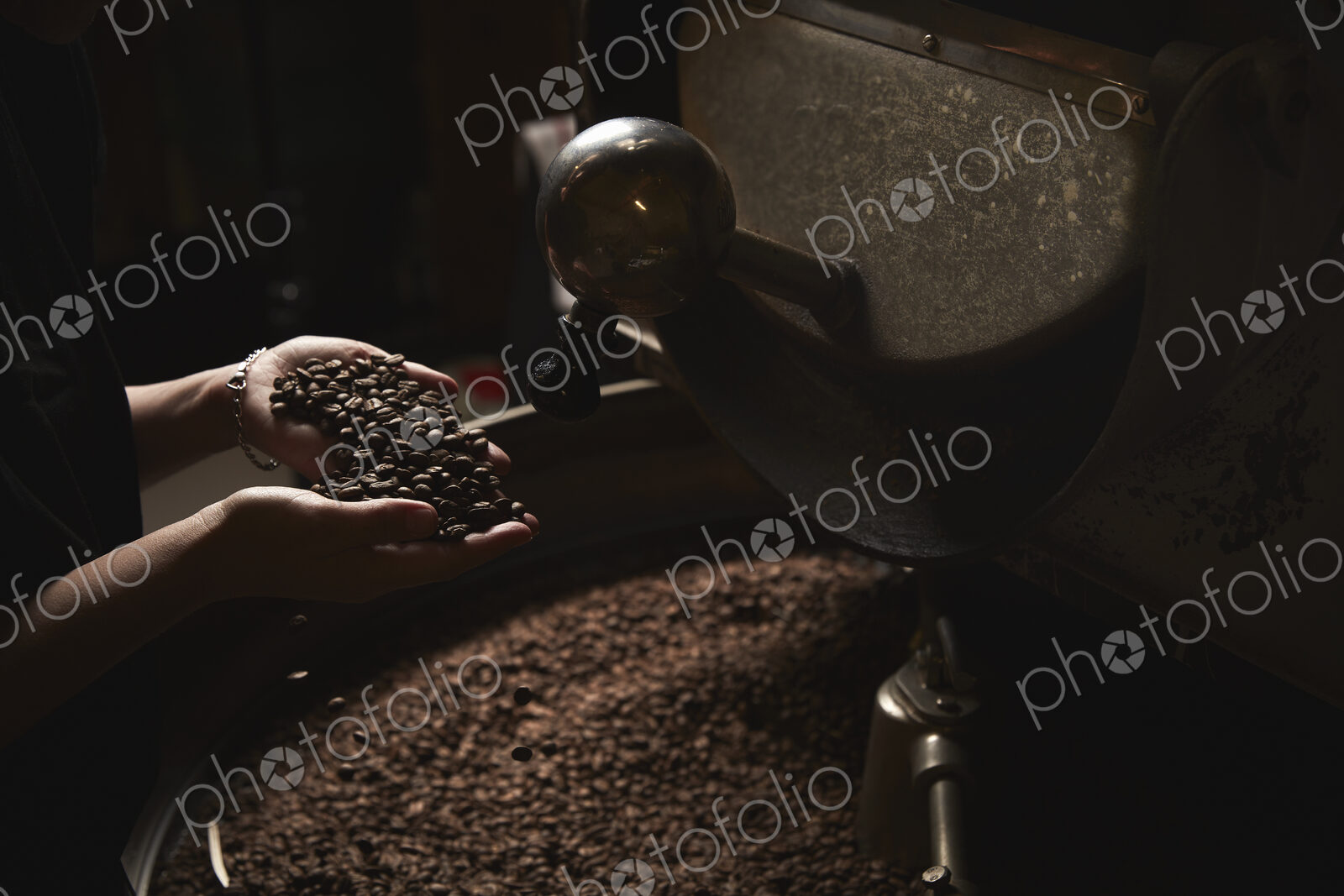 A coffee shop. A person holding a handful of fresh roasted beans above a metal drum…