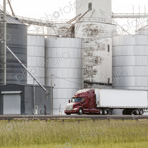 A commercial truck driving past grain elevators in the farm country of eastern… A commercial truck driving past grain elevators in the farm country of eastern…
