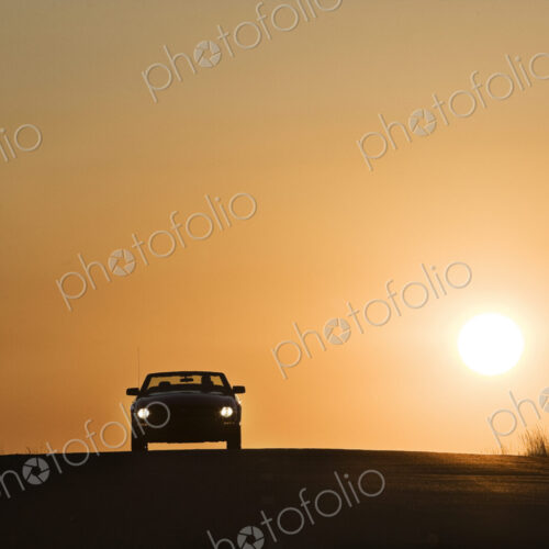 A convertible sports car on a country highway at sunset.
