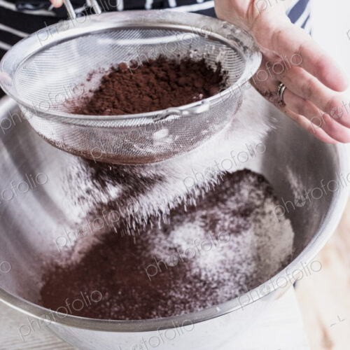 A cook sieving cocoa powder into a metal bowl of flour and cake ingredients. A cook sieving cocoa powder into a metal bowl of flour and cake ingredients.