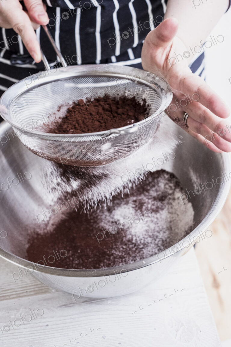 A cook sieving cocoa powder into a metal bowl of flour and cake ingredients.