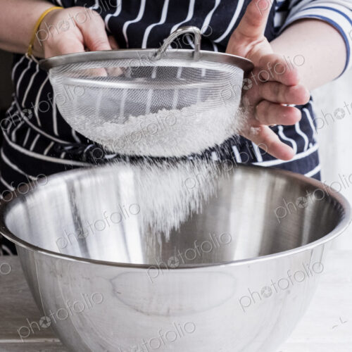 A cook sieving ingredients through a sieve, flour into a metal bowl. A cook sieving ingredients through a sieve, flour into a metal bowl.