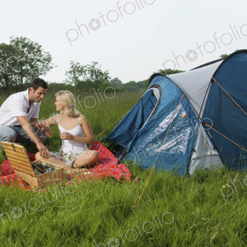 A couple, a man and woman seated having a picnic. A blue tent. Camping.