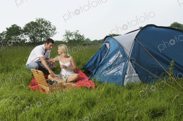 A couple, a man and woman seated having a picnic. A blue tent. Camping.