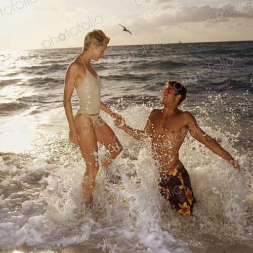 A couple in the sea, leaping in the surf at sunset. A man kneeling in the water.
