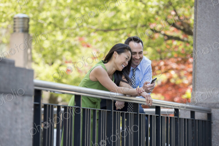 A couple, man and woman checking a smart phone, under the shade of trees in the park.