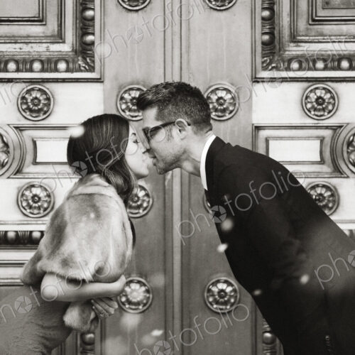A couple, man and woman kissing in front of the large decorated doors of a city building. A couple, man and woman kissing in front of the large decorated doors of a city building.