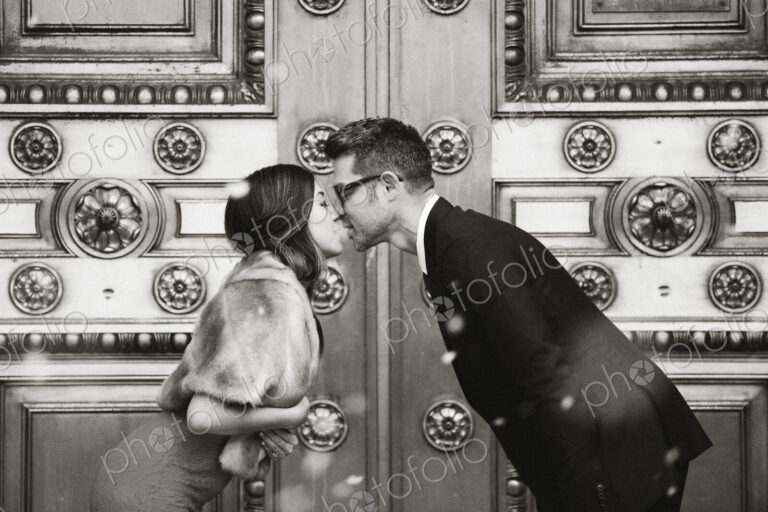 A couple, man and woman kissing in front of the large decorated doors of a city building.