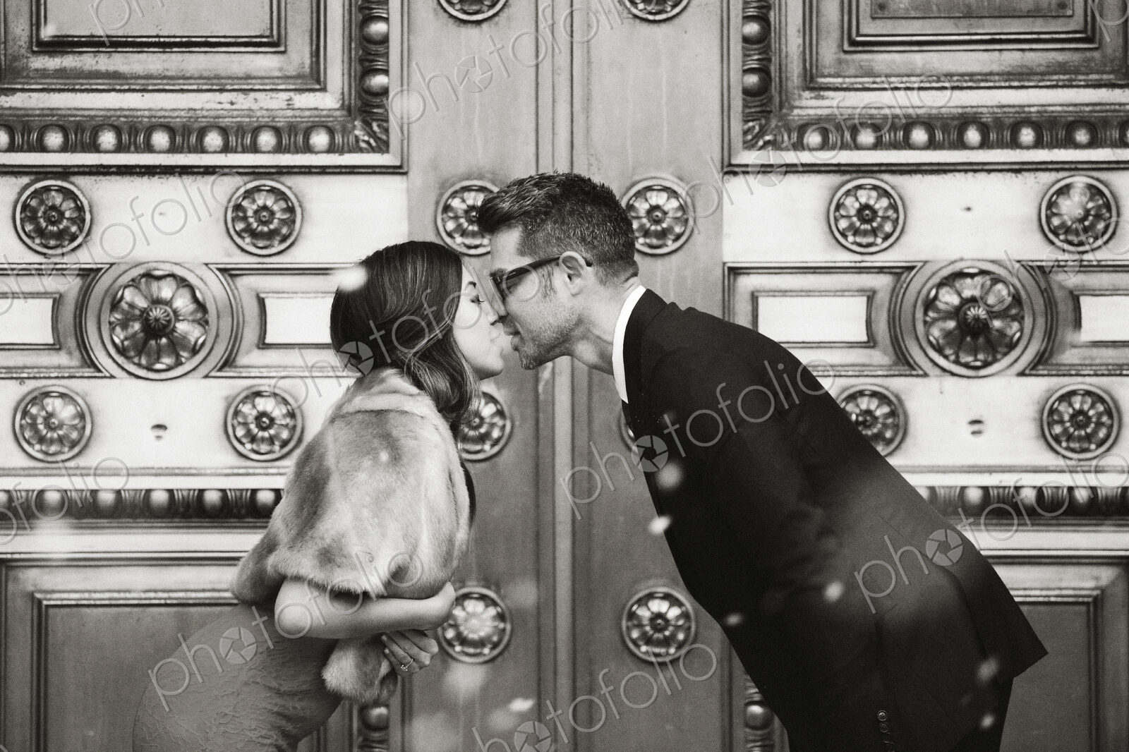 A couple, man and woman kissing in front of the large decorated doors of a city building.