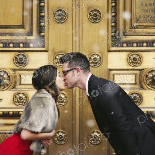 A couple, man and woman kissing in front of the large decorated doors of a city building.