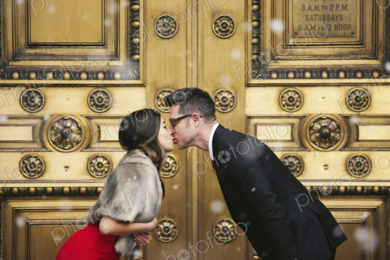 A couple, man and woman kissing in front of the large decorated doors of a city building.