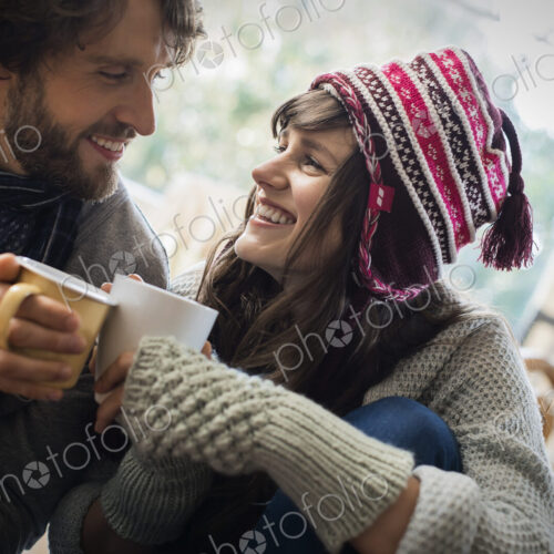 A couple, man and woman sitting close together warming up with their hands around mugs.