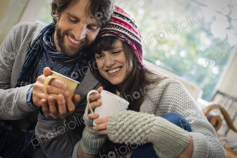 A couple, man and woman sitting close together warming up with their hands around mugs.