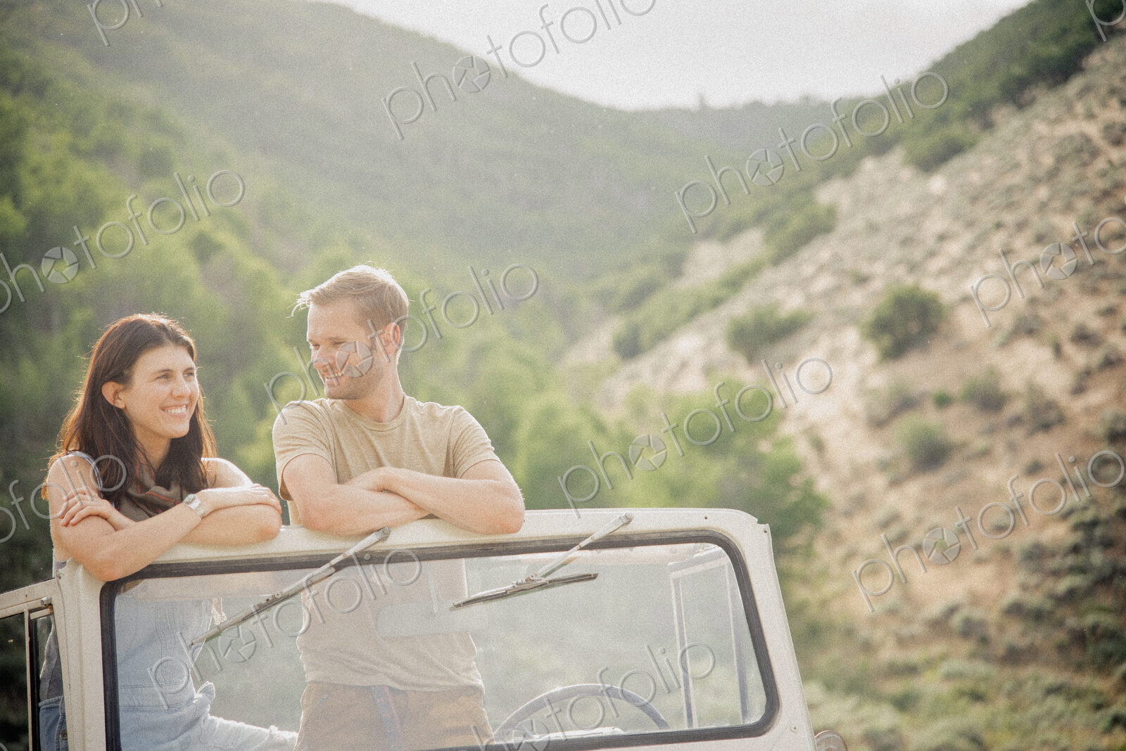 A couple on a road trip in the mountains standing side by side in a jeep looking around.