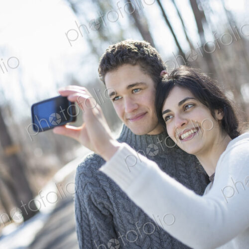 A couple outdoors on a snowy day. Woman holding a camera phone, taking photographs at… A couple outdoors on a snowy day. Woman holding a camera phone, taking photographs at…