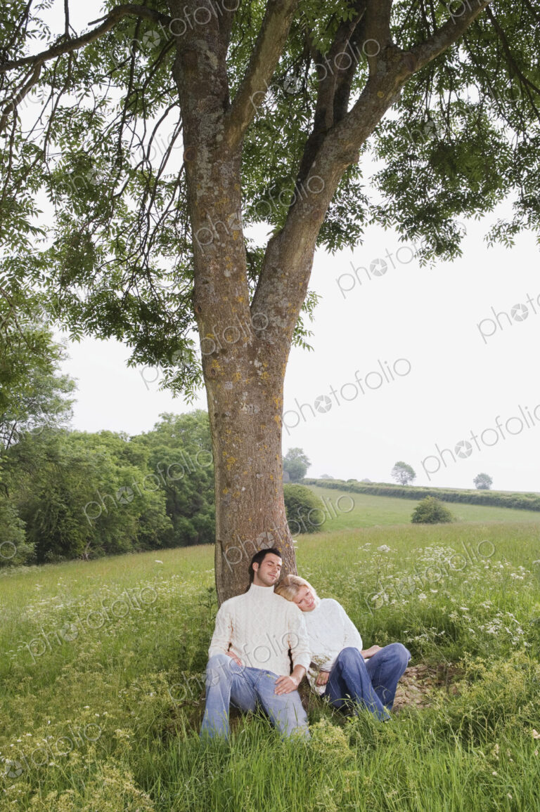 A couple sitting leaning against a tree in a meadow.