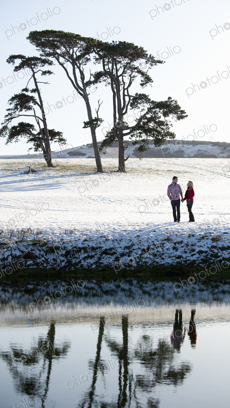 A couple standing in a snow-covered field by a river, holding hands.