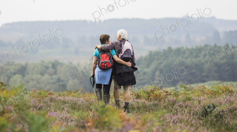A couple standing on a path looking at the view over wooded hills, with arms around…