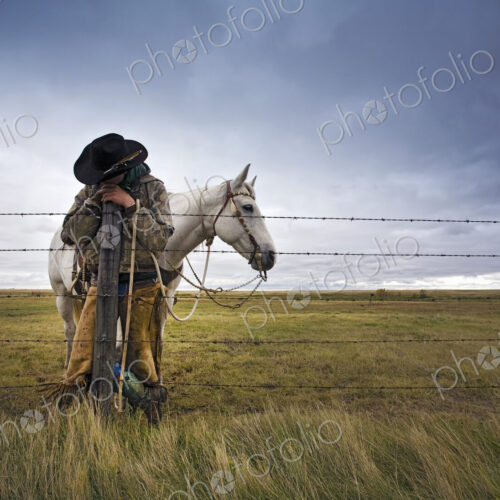 A cowboy standing leaning on a fence post on the range. A grey horse behind him. A cowboy standing leaning on a fence post on the range. A grey horse behind him.