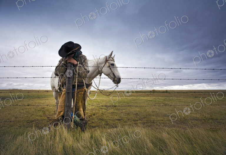 A cowboy standing leaning on a fence post on the range. A grey horse behind him.