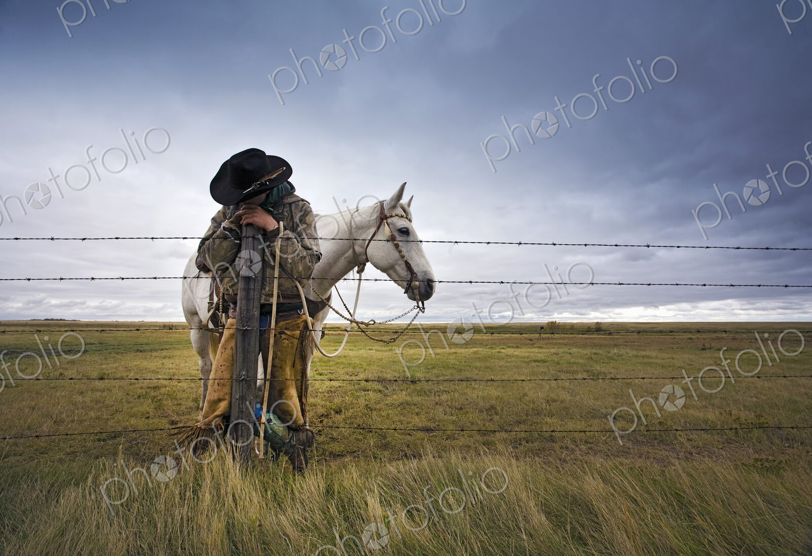 A cowboy standing leaning on a fence post on the range. A grey horse behind him.