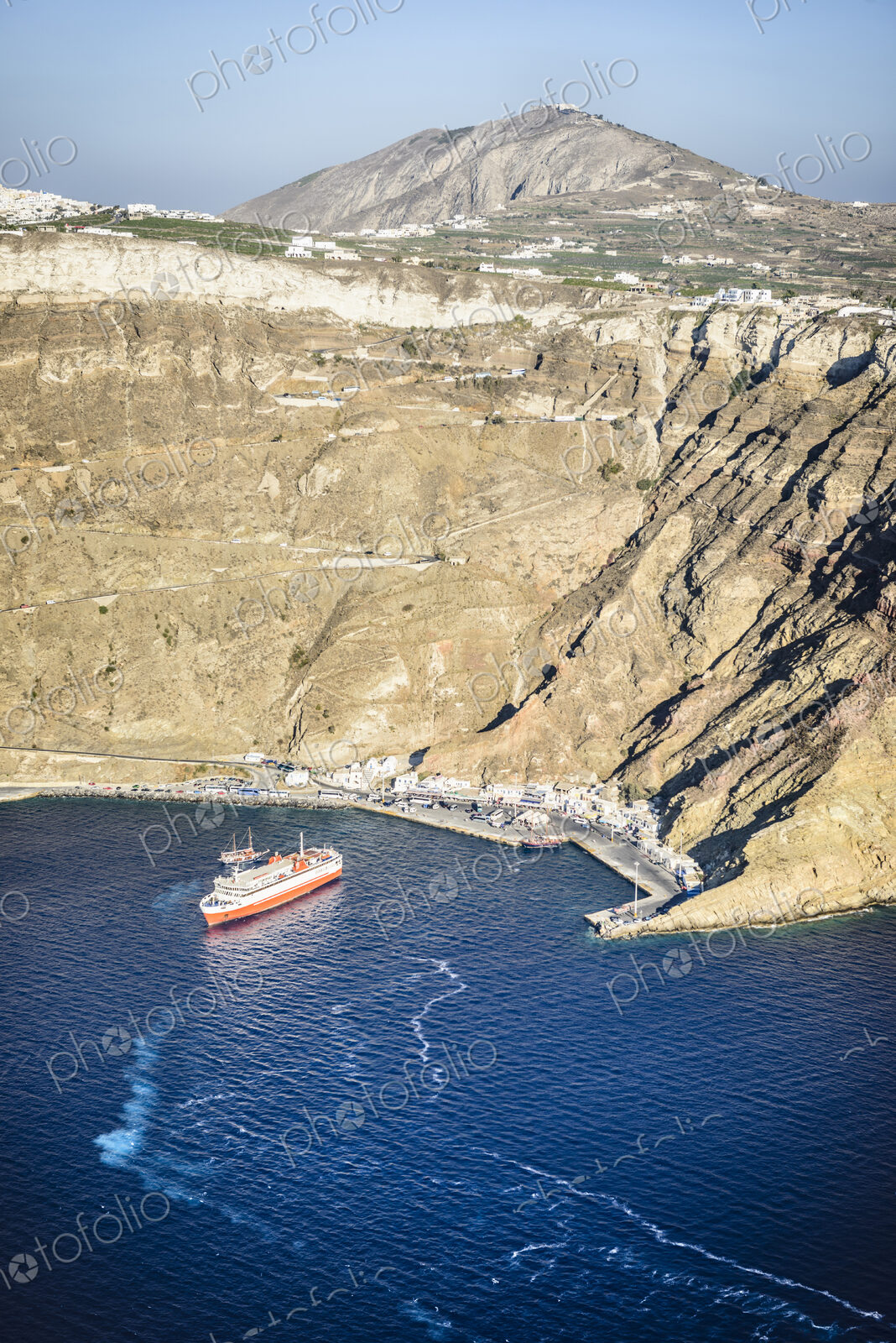 A cruise ship approaching the harbour of Oia on the coastline at the base of a steep…