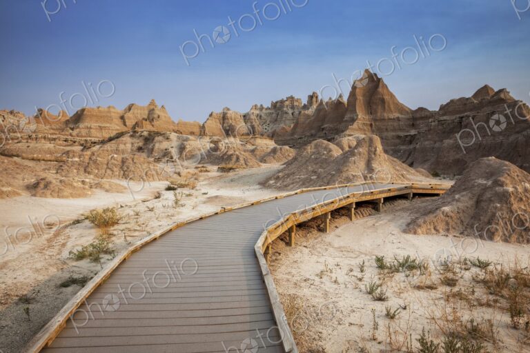 A curving boardwalk leads into eroded buttes and pinnacles under a clear blue sky in…