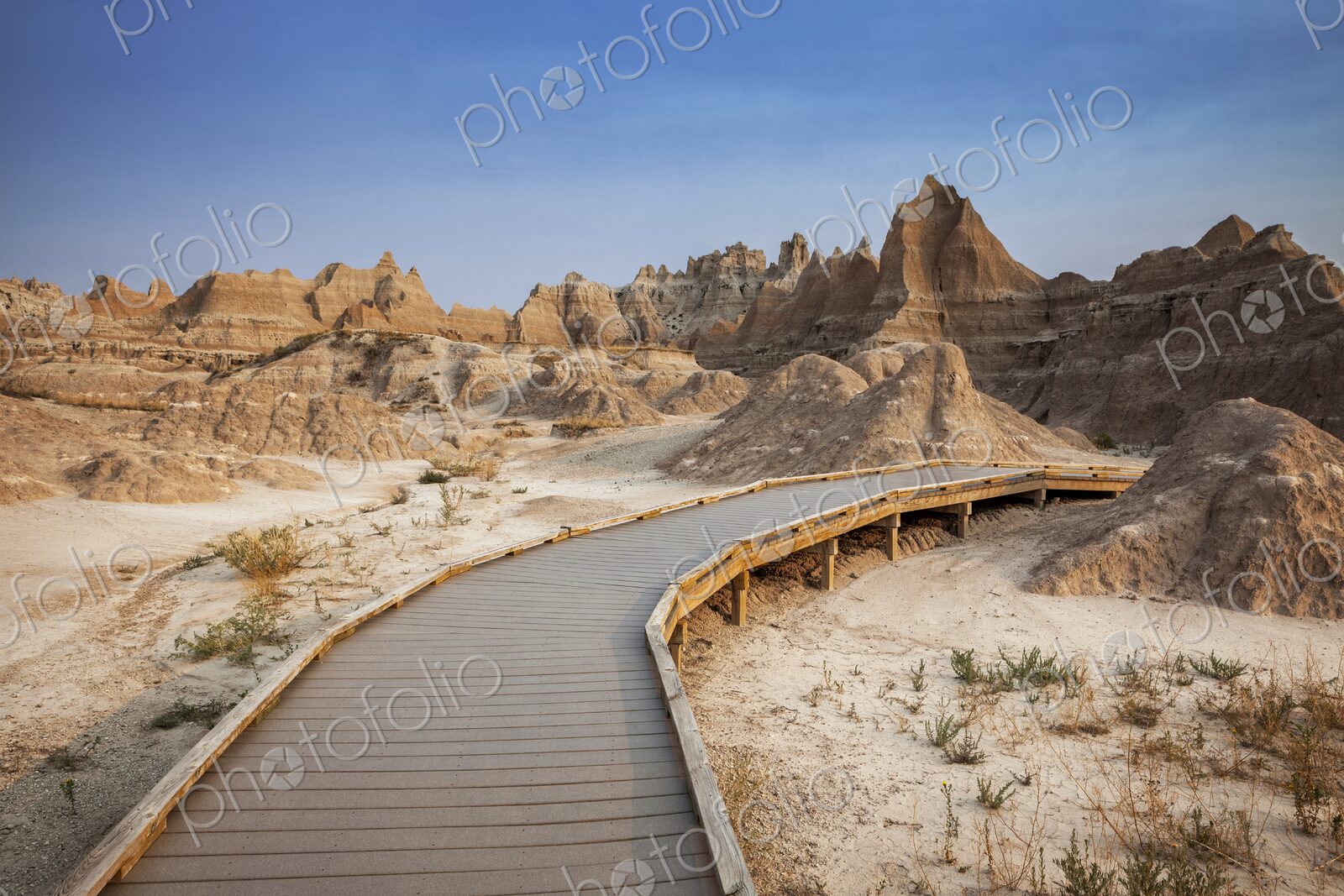 A curving boardwalk leads into eroded buttes and pinnacles under a clear blue sky in…