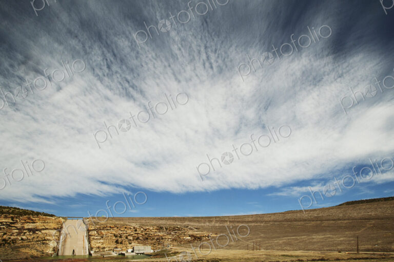 A dam wall and sluice overflow into a lake, with a blue sky and streaked cloud pattern.