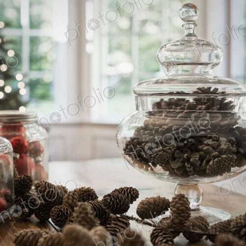 A decorative glass jar of pine cones and red glass baubles on a table top. Christmas…