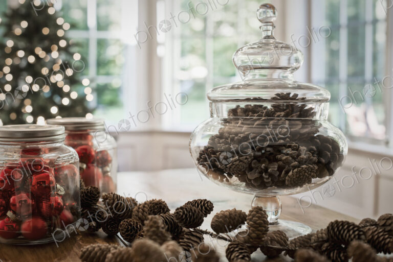 A decorative glass jar of pine cones and red glass baubles on a table top. Christmas…