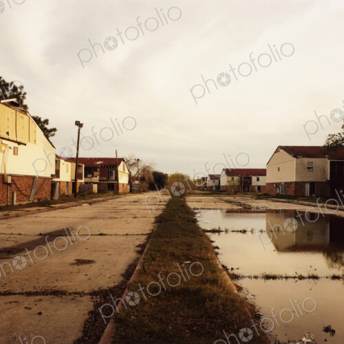 A deserted road, with cracked paving and puddles, and industrial buldings.