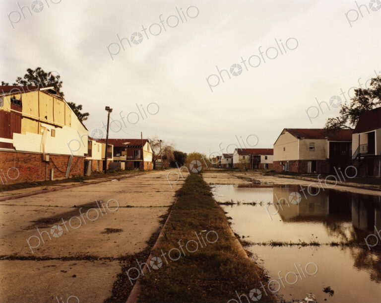 A deserted road, with cracked paving and puddles, and industrial buldings.