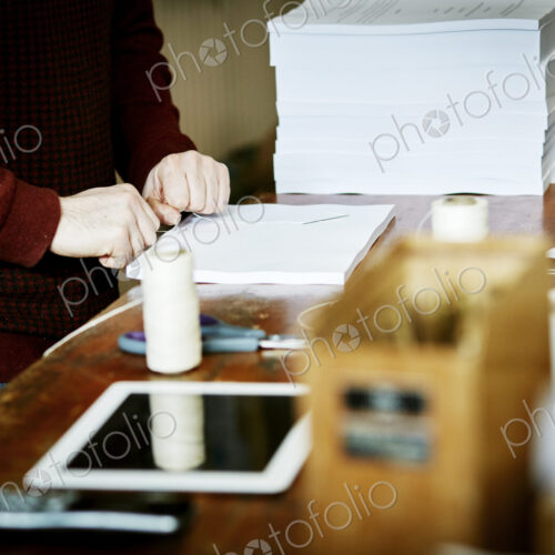 A digital tablet on a workbench, and a man handstitching a pile of paper pages.