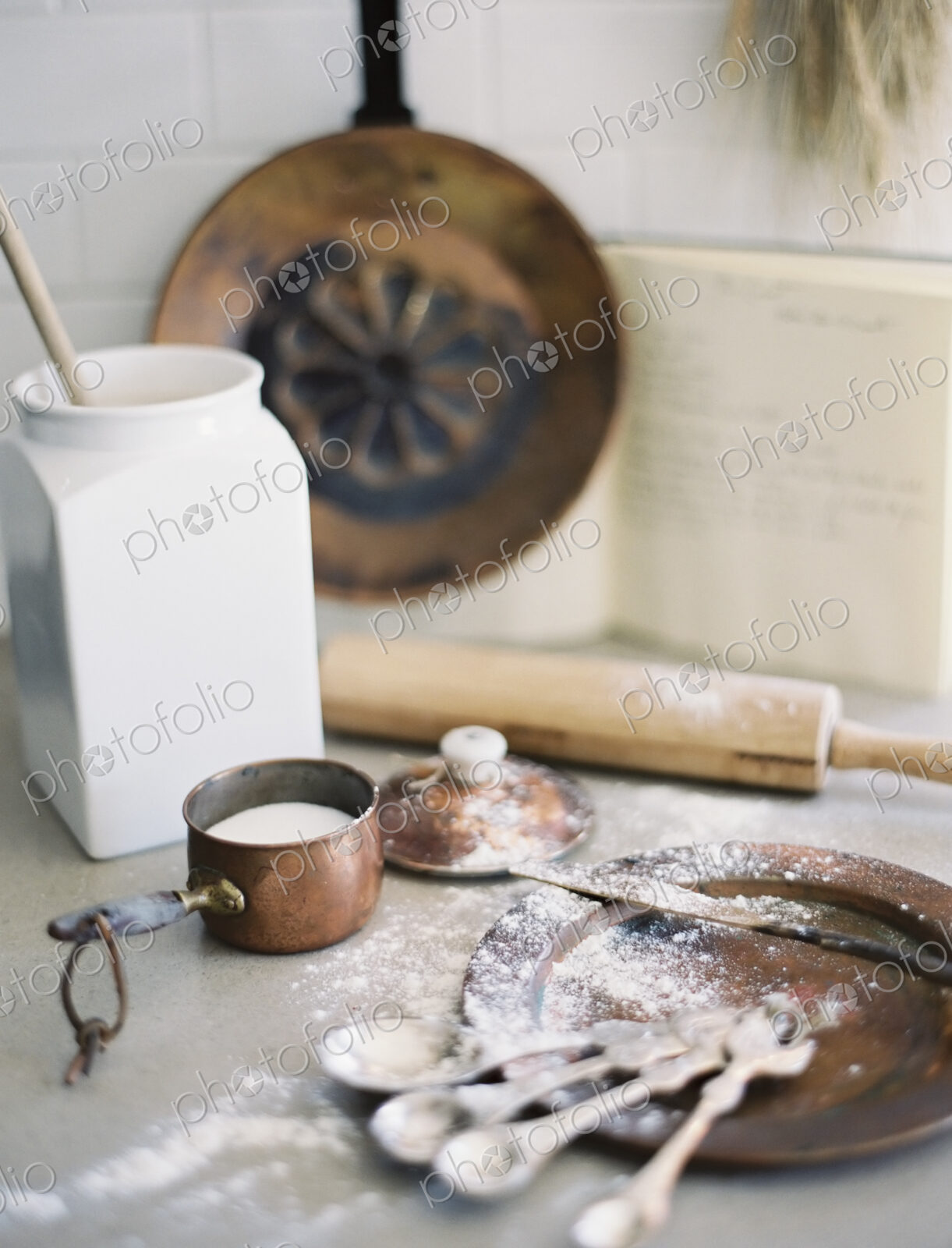 A domestic kitchen table. A large group of kitchen utensils, spoons, a pan and a…