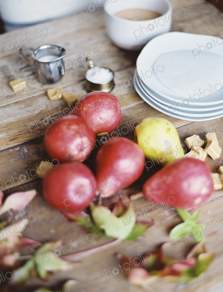 A domestic kitchen tabletop. A small group of fresh organic pears and a stack of white…