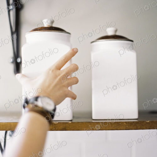 A domestic kitchen. A person reaching up to two storage jars on a shelf. A domestic kitchen. A person reaching up to two storage jars on a shelf.