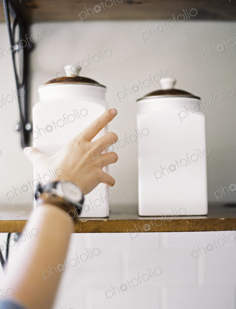 A domestic kitchen. A person reaching up to two storage jars on a shelf.