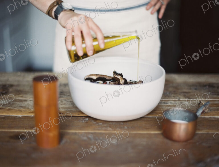 A domestic kitchen. A woman wearing an apron, dressing a fresh salad of vegetables in…
