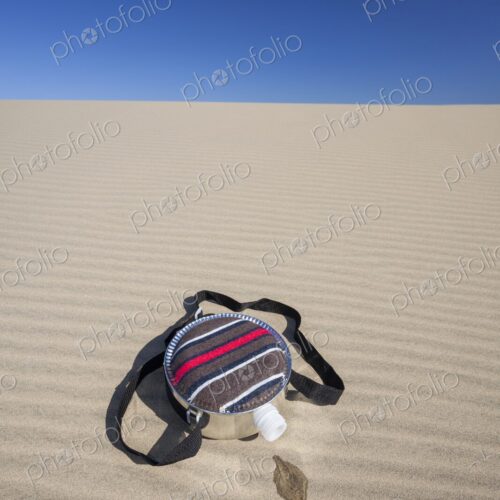 A fabric-covered canteen rests on rippled sand dunes with a small puddle of spilled…