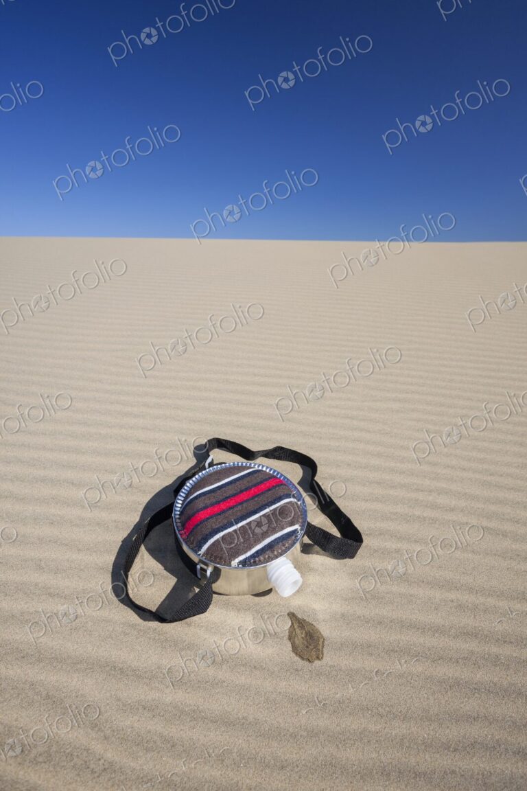 A fabric-covered canteen rests on rippled sand dunes with a small puddle of spilled…